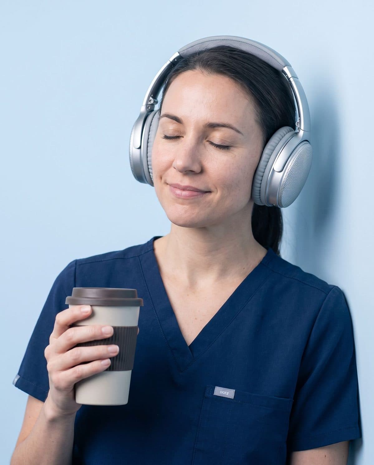 Nursing student in scrubs with headphones on and eyes closed, coffee in hand.